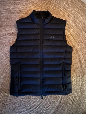 Svart dunväst från j.lindberg - Snygg mörkblå dunväst från J.lindberg med quiltad design. Västen har dragkedja framtill och två sidofickor. Perfekt för lager-på-lager och håller dig varm. Orginal pris ca 2000kr. Storlek xs men passar S