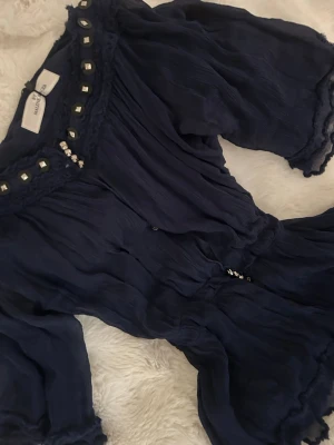 Unik Mörkblå blus med detaljer från By Malene Birger - Supersnygg unik mörkblå blus från By Malene Birger med dekorativa silverfärgade nitar längs ringningen. Blusen har trekvartsärm, lätt rynkad struktur och dragsko i midjan för en skön passform. Materialet är tunt och luftigt, perfekt för en trendig look.