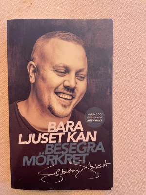 Bara ljuset kan besegra mörkret - En bok med svart omslag och porträttbild, titeln 'Bara ljuset kan besegra mörkret' och författarens signatur på framsidan. Boken handlar om kampen mellan ljus och mörker, och om att hitta hopp och förändring i livet.
