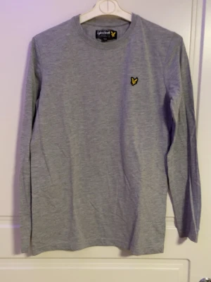 Grå långärmad tröja Lyle & Scott - Snygg grå långärmad tröja från Lyle & Scott med rund halsringning och den klassiska gula loggan broderad på bröstet. Tillverkad i mjukt bomullsmaterial som känns skönt mot huden. Perfekt basplagg för en clean och enkel stil.