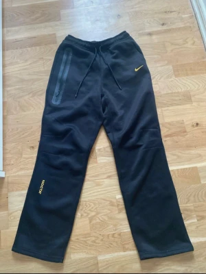 Svarta mjukisbyxor från Nike NOCTA - Säljer ett par svarta mjukisbyxor från Nike NOCTA med gul logga och text. Byxorna har snörning i midjan, dragkedjeförsedd ficka och raka ben. Perfekta för chill eller streetwear-stil.