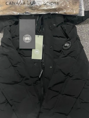 Svart pufferjacka från Canada Goose - Säljer en svart pufferjacka från Canada Goose med huva och diskret logga på ärmen. Jackan har dragkedja framtill, breda vadderade paneler och en klassisk look. Perfekt för kalla dagar och riktigt snygg till streetwear-stilen.