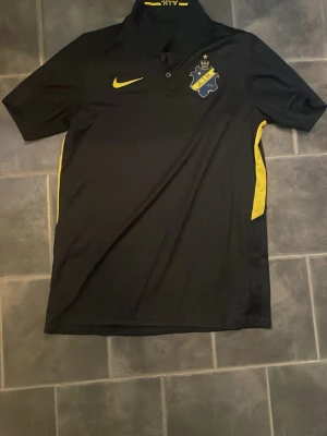 Svart AIK pikétröja från Nike - Snygg svart pikétröja från Nike med AIK:s klubbmärke på bröstet och gula detaljer längs sidorna. Tröjan har korta ärmar, krage med knappar och är tillverkad i ett sportigt funktionsmaterial. Perfekt för dig som vill representera AIK både på och utanför planen.