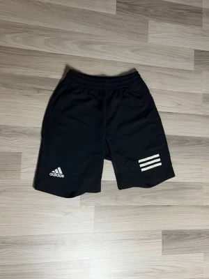 Svarta Adidas shorts - Svarta shorts från Adidas med elastisk midja och klassiska tre vita ränder på ena benet. Passar bra om du är runt 170-180cm. Tillverkade i lätt och bekvämt syntetmaterial, perfekta för sport eller chill. Enkel och stilren design.