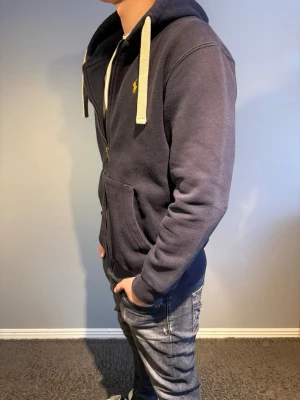 Ralph Lauren hoodie - Mörkblå hoodie från Ralph Lauren med dragkedja, vita snören och den klassiska gula loggan på bröstet. Den har storlek L men passar mer som M. Modellen är 184.