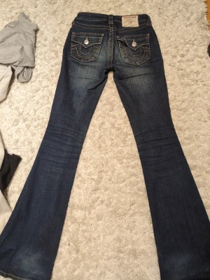 True Religion mörkblå bootcut jeans - Snygga mörkblå jeans från True Religion med klassiska kontrastsömmar och stora fickor med vita knappar bak. Modellen har bootcut-ben, vilket ger en riktigt cool siluett. Jeansen är tillverkade i jeansmaterial med orange detaljer vid fickorna. Meddela om mått