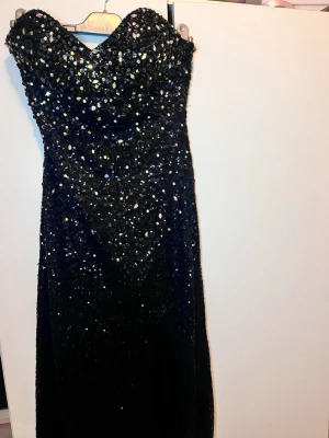 Svart glittrig långklänning - Supersnygg svart långklänning med massor av paljetter och glittriga detaljer. Axelbandslös modell med figurnära passform och hjärtformad urringning. Perfekt för bal eller festkvällar när du vill glänsa extra mycket.