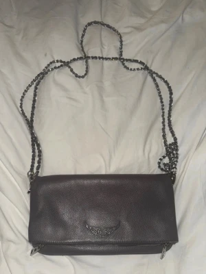 Zadig Voltaire väska - Snygg mörkbrun axelväska i skinn med silverfärgade kedjeremmar och dragkedjedetaljer. Väskan har en stilren design med en liten metallogo framtill och extra fack med dragkedja på baksidan. Perfekt för att bära det viktigaste.