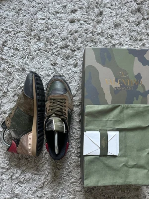 Valentino Rockrunner sneakers camo - Säljer ett par Valentino Rockrunner sneakers i camo-mönster med gröna, bruna och svarta toner. Skorna har detaljer i mocka och skinn, coola nitar vid hälen och röd accent baktill. Klassisk snörning och platt sula. Kommer ej med originalkartong och dustbag. Kan dock skicka med vid förhandling 