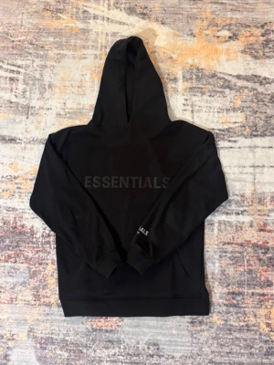 Svart Essentials hoodie - Säljer en svart hoodie från Essentials med diskret logga på bröstet och textdetalj på ärmen. Klassisk passform med stor huva och ribbade muddar. Perfekt för dig som gillar stilrena och bekväma plagg. Materialet känns mjukt och skönt mot huden.