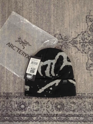 Svart mössa från Arc'teryx - Snygg svart mössa från Arc'teryx med grått grafiskt mönster på utsidan och diskret logga. Mössan är vändbar med en helsvart sida och orange insida. Tillverkad i mjukt, stretchigt material som sitter skönt på huvudet.