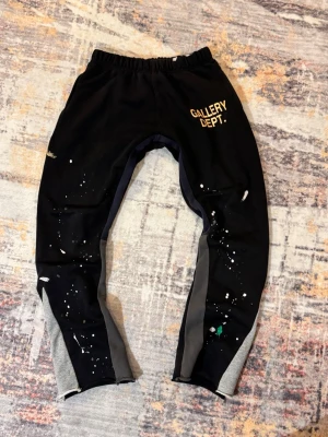 Svarta mjukisbyxor Gallery Dept. - Unika svarta mjukisbyxor från Gallery Dept. med grå paneler längs benen och färgstänk som ger en kreativ vibe. Byxorna har resår i midjan och tryckt logga på vänster lår. Perfekta för dig som gillar streetwear och vill sticka ut.