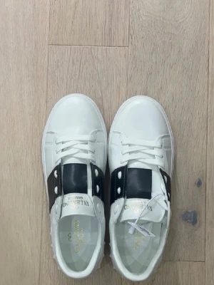 Valentino Open vita sneakers - Snygga vita Valentino Open sneakers med bred svart rem över vristen och vita nitar på sidan. Klassisk rund tå, snörning och exklusiv känsla. Tillverkade i skinn med subtila detaljer och guldfärgad logga på plösen. Finns i storlek 43
