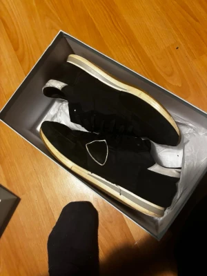 Philippe model  - Säljer ett par svarta sneakers med vit och beige sula. Skorna har snörning och en tydlig vit detalj på sidan. De har en klassisk låg profil och är tillverkade i en mix av skinn och syntetmaterial. Hål som ni kan se på bilden 