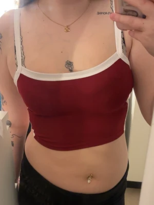 Röd croppad topp med vita band - Säljer en croppad röd topp med vita axelband och kantdetaljer. Toppen har en tight passform och är ärmlös, perfekt för varma dagar. Materialet känns mjukt och stretchit.