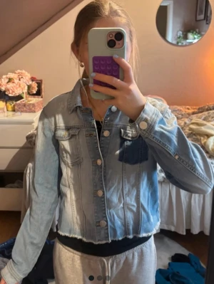 Ljusblå croppad jeansjacka - Trendig ljusblå jeansjacka med croppad passform och rå kant nertill. Klassisk design med två bröstfickor och knappar framtill. Perfekt för att lyfta vilken outfit som helst.