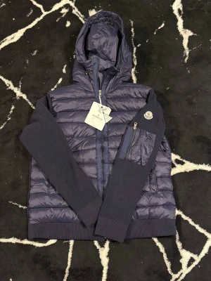 Mörkblå dunjacka från Moncler - Snygg mörkblå dunjacka från Moncler med quiltad front och huva. Jackan har ribbade stickade ärmar och detaljer, dragkedja framtill samt en ficka med dragkedja på ärmen. Perfekt för kyliga dagar och har en modern, sportig look.