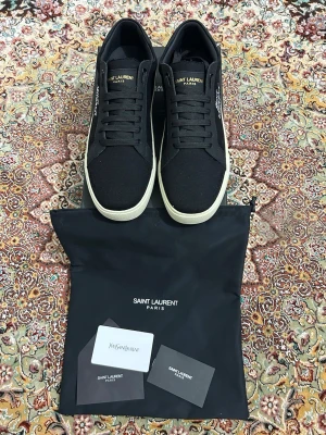 Svarta Saint Laurent sneakers - Snygga svarta sneakers från Saint Laurent Paris med vit sula och klassisk logga på sidan. Skorna har snörning och är tillverkade i canvas med detaljer i skinn. Perfekta för en stilren och trendig look.