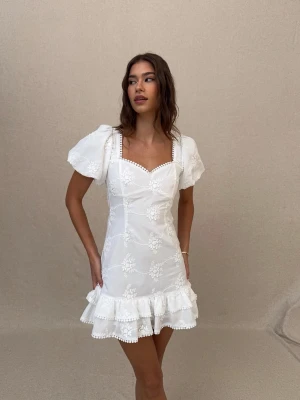 Vit studentklänning med puffärm och rosett - Superfin studentklänning med broderade blommor, puffärmar och volangkant. Öppen rygg med stor rosett och söta spetsdetaljer vid halsringningen. Aldrig använd! Prislappen sitter fortfarande kvar.