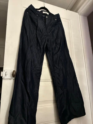 Mörkblå wide leg jeans från Kappahl, storlek 38 - Säljer ett par mörkblå jeans från Kappahl med breda ben och hög midja. Klassisk femficksmodell med knapp och dragkedja framtill. Snygg denim i 100% bomull, perfekt för en trendig och avslappnad stil.