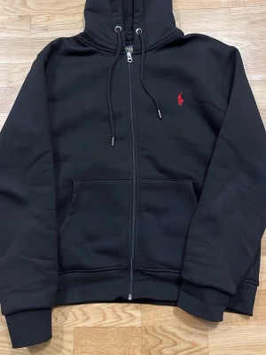 Svart hoodie från Polo Ralph Lauren - Svart hoodie med dragkedja från Polo Ralph Lauren. Klassisk design med luva och dragsnören. Helt ny nästan o använd 10/10 skick. 