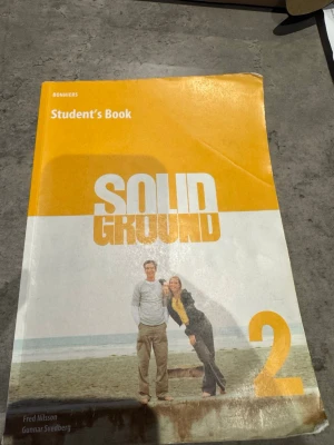 Solid Ground 2 Student's Book - Engelsk kursbok med gul och vit framsida, titeln 'Solid Ground 2'. Omslaget visar två personer på en strand. Författare: Fred Nilsson och Gunnar Svedberg. Perfekt för gymnasieelever som läser engelska.