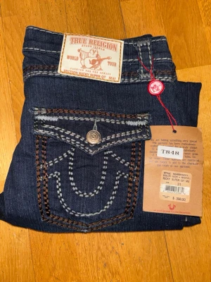 True Religion Ricky Super QT Unisex – Made in USA – Medium (W31) - Helt oanvända True Religion Ricky Super QT jeans, nya med originalprislappar kvar. Rak modell som fungerar bra som unisex. Tillverkade i USA av 100 % bomull.  Storlek Medium (W31). Mått (mätta plant): midja 40 cm, innerben 76 cm, benlängd fram 97 cm, benlängd bak 102 cm.