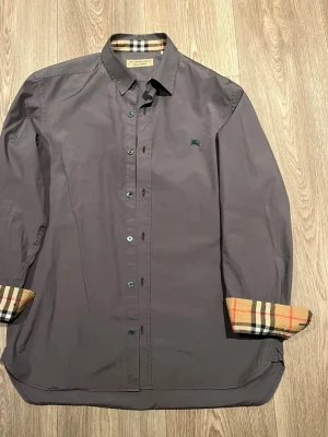 Grå Burberry skjorta med rutiga detaljer - Säljer en tvärfet grå skjorta från Burberry med klassiska rutiga detaljer på insidan av kragen och manschetterna. Skjortan har knappar framtill och en liten broderad logga på bröstet. Skjortan är i storlek M. Tveka inte på att höra av dig vid frågor! 