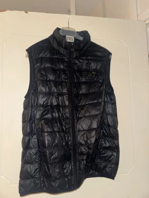Marinblå dunväst från EA7 Emporio Armani - Snygg svart/marinblå dunväst från EA7 Emporio Armani med glansig finish och diskret logga på bröstet. Västen har hög krage, dragkedja framtill och två sidofickor. Perfekt för lager på lager och ger en sportig vibe till din outfit.