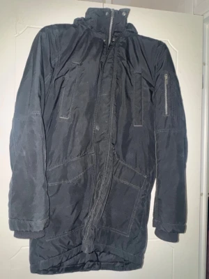 Svart parkasjacka från H&M - Säljer en svart parkasjacka från H&M med hög krage och flera praktiska fickor, inklusive en på ärmen. Jackan har dragkedja framtill och ribbade muddar vid ärmsluten. Perfekt för kyliga dagar och enkel att matcha med olika outfits. Behövs tvättas.