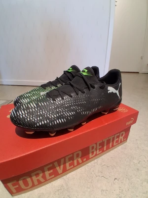 Svarta Puma fotbollsskor  - Säljer ett par svarta fotbollsskor från Puma med vitt och grönt mönster. Skorna har snörning och är designade för att ge bra grepp på planen. Puma-loggan syns tydligt på sidan och tungan. Perfekta för dig som vill sticka ut på fotbollsplanen.