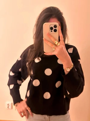 Svart stickad tröja med vita prickar - Säljer en svart stickad tröja med stora vita prickar. Tröjan har en avslappnad passform, rund halsringning och långa ärmar. Perfekt för dig som gillar lekfulla mönster och vill ha något som sticker ut i garderoben.