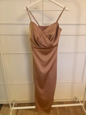 Champagnefärgad långklänning i satin - Elegant långklänning i rosa satin med smala axelband. Klänningen har en figurnära passform och en mjuk glansig yta som ger en lyxig känsla. Perfekt för dig som vill sticka ut med en stilren look.