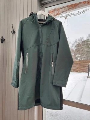 Mörkgrön regnjacka med huva - Grön fleecefodrad softshellkappa med dragkedja fram och tre fickor med dragkedja. Jackan har en huva och svarta detaljer på sidorna. Färgen heter Mossgreen.  Kappan är i mycket bra skick, ser knappt använd ut och kostar 1499 kr i handeln.