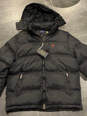 Svart dunjacka från Polo Ralph Lauren - Snygg svart dunjacka från Polo Ralph Lauren med röd logga på bröstet. Jackan har huva, dragkedja framtill och två dragkedjeförsedda fickor. Perfekt för kalla dagar och riktigt stilren look. Det är S på storleken men skulle mer säga att de är M! 