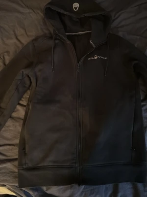 Svart hoodie från Sail Racing - Säljer en svart hoodie från Sail Racing med dragkedja framtill och två sidofickor. Hoodien har justerbar huva med snören och diskret logga på bröstet samt på huvan. Materialet känns mjukt och slitstarkt, perfekt för en avslappnad stil.