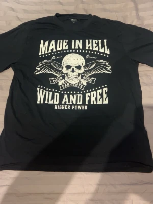 Svart t-shirt med dödskalletryck - Cool svart t-shirt från INEXTENSO med stort vitt tryck framtill. Texten 'MADE IN HELL' och 'WILD AND FREE' tillsammans med en dödskalle och vingar ger en edgy vibe. Klassisk passform och rund halsringning. Perfekt för dig som gillar statement-plagg.