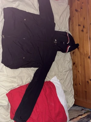 Moncler Windbreaker - Snygg svart vindjacka från Moncler med huva och dragkedja framtill. Jackan har Moncler-logga på ärmen och detaljer i rött och vitt på huvan. Perfekt för dig som vill ha en stilren och sportig look. Sitter som en S me passar M bra! Är 185 och sitter perfekt!