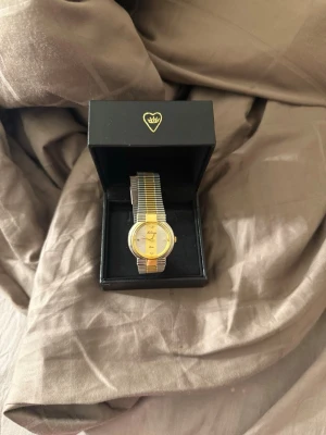 La Strada guldfärgad klocka - Snygg klocka från La Strada med rund urtavla i guld och silver. Armbandet är i metall med guldfärgade och silverfärgade ränder. Urtavlan har eleganta detaljer och små stenar vid siffrorna. Perfekt accessoar för dig som gillar stilrena och klassiska klockor.(inget batteri)