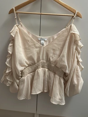 Beige volang topp från NLY Trend - Supersöt beige topp från NLY Trend med tunna axelband, v-ringning och volangdetaljer längs ärmarna. Toppen har smock i midjan för en snygg passform och är tillverkad i ett lätt, flowy material. Perfekt för dig som gillar romantisk stil!
