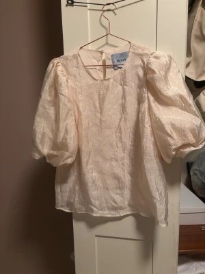 Krämfärgad puffärmblus från Minus - Säljer en krämfärgad blus från Minus med puffiga korta ärmar och rund halsringning. Blusen har en lätt glansig struktur och är tillverkad i ett tunt, skirt material som ger en elegant look. Perfekt för dig som gillar trendiga detaljer och volym.