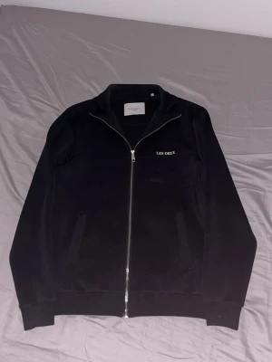 Svart ziptröja från Les Deux - Säljer en svart ziptröja från Les Deux med stilren design och diskret logga på bröstet. Tröjan har hel dragkedja, ståkrage och två fickor framtill. Materialet känns mjukt och har en lätt glansig finish, perfekt för en clean och modern look.