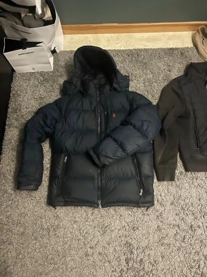 Mörkblå dunjacka från Polo Ralph Lauren - Denna mörkblåa dunjacka från ralph lauren är i högsta kvalité och har blivit väl behållen. Köpt för 5499 och säljs för ett extremt bra pris på 2500. Har kvar en broschyr som man får med när man köper jackan och kan även skicka fler bilder. Det saknar en liten metall bit i slutet på drag kedjan vid luvan vilket gör att den är lite bökig att få på men annars funkar allt fint. 