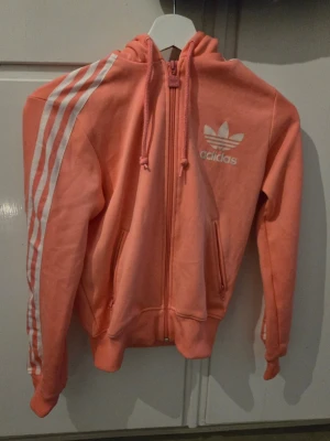 Rosa retro vintage hoodie från Adidas med dragkedja - Säljer en snygg rosa retro hoodie från Adidas med vit logga och klassiska tre ränder på ena ärmen. Hoodien har dragkedja framtill, två fickor och justerbar huva med snören. Perfekt för dig som gillar sportig och avslappnad stil.
