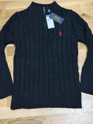 Svart kabelstickad tröja Polo Ralph Lauren - Kabelstickad svart tröja från Polo Ralph Lauren med halv dragkedja och ribbade muddar. Klassisk röd logga broderad på bröstet. Tröjan har hög krage och är tillverkad i mjukt material som passar perfekt till höst och vinter.