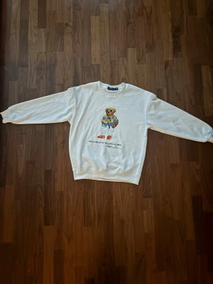 Vit sweatshirt Polo Bear Ralph Lauren - Vit sweatshirt från Ralph Lauren med ikoniska Polo Bear-trycket på bröstet. Tröjan har rund hals, långa ärmar och ribbade muddar. Materialet ser ut att vara mjukt bomullstyg. Perfekt för dig som gillar klassisk streetwear med en twist.