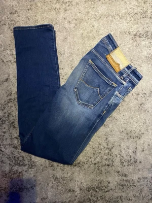 Blå jeans från Jacob Cohën - Snygga blå jeans från Jacob Cohen med klassisk femficksmodell och unikt läderpatch bak i beige och brunt. Jeansen har raka ben och kontrastsömmar som ger en stilren look. Det enda att notera är en liten röd färgfläck på höger ben vid ankeln ungefär. Fläcken syns inte vid användning som tur är men ändå värt att notera. Hör gärna av dig vid frågor!