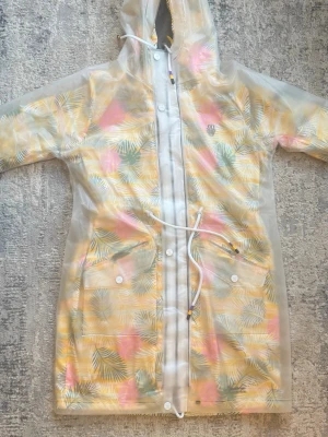 Mönstrad regnjacka från Navahoo - Transparant regnjacka från Navahoo med tropiskt bladmönster i gult, rosa och grönt. Jackan har huva, dragsko i midjan och stängs med både dragkedja och knappar. Perfekt för regniga dagar när du vill sticka ut lite extra.