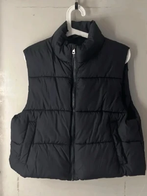 Svart dunväst  - Säljer en svart dunväst från lager 157. Västen har hög krage, dragkedja framtill och två sidofickor. Klassisk quiltad design som är både stilren och enkel att matcha till olika outfits. Perfekt för lager på lager under kyliga dagar.