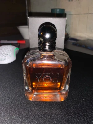 Stronger With You parfym från Armani - 90ml kvarSäljer efter som den inte används längre. Stronger With You från Emporio Armani är en elegant parfym i en genomskinlig, fyrkantig glasflaska med rund svart kork. Doften har en varm bärnstensfärgad vätska och flaskan har stilren text i vitt framtill. Perfekt för dig som gillar exklusiva dofter.